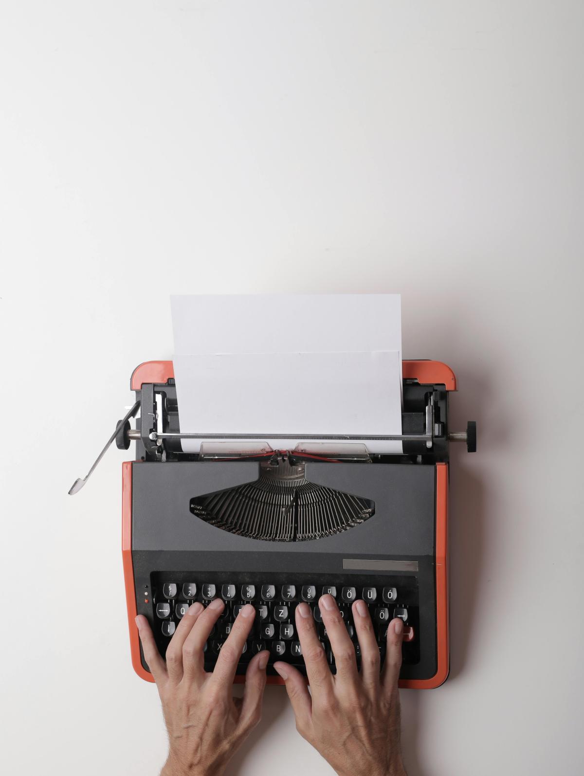 typewriter with two hands poised to type on a blank piece of paper